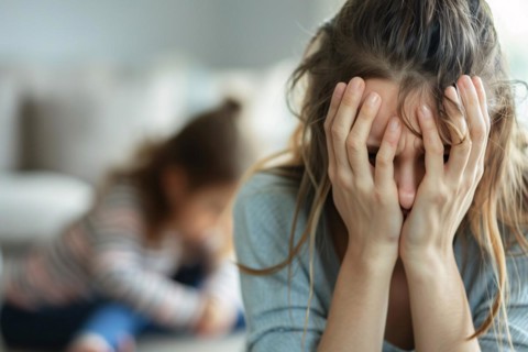Stressed and frustrated young mother hiding face in hands with toddler child in blurry background.