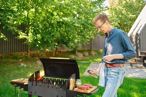 Middle-aged man sprinkles raw steak with oil from bottle while cooking at backyard