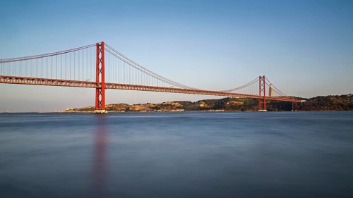 Serene dusk view of a red suspension bridge over calm blue water with reflections, Lissabon, 25 de