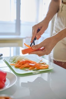 An unrecognizable woman peeling carrots on a green cutting board in a sunlit kitchen. Slices of
