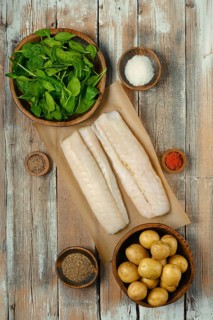Fresh fish fillets on parchment paper surrounded by bowls of spinach, potatoes, and various spices,