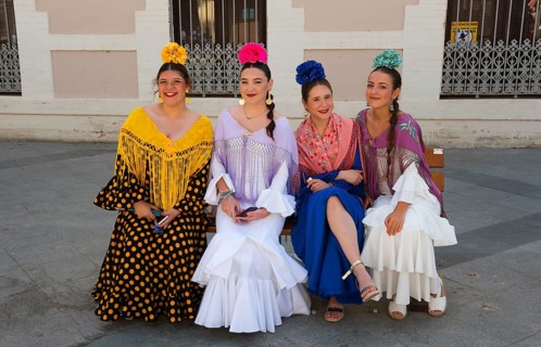 Four woman in colourful flamenco dresses and flowers in their hair sit on a bench on a street in