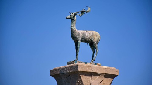 Bronze deer statue on a pedestal in front of a clear blue sky, Mandraki harbour, harbour area,