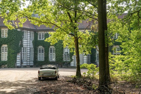 Vintage car parked in front of a historic, ivy-covered stately building surrounded by trees in the