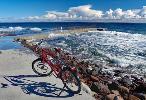A red bicycle stands on the shore with a view of the rough sea and thick clouds in the blue sky,