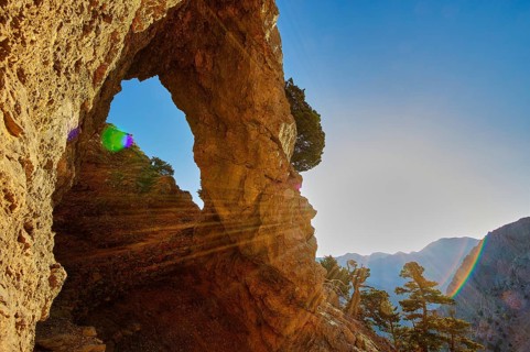 A rocky arch with sunbeams, surrounded by trees and mountains under a clear sky, path to the summit