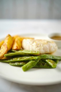 A tender fish fillet alongside fresh green beans and golden roasted potatoes. A small bowl of sauce