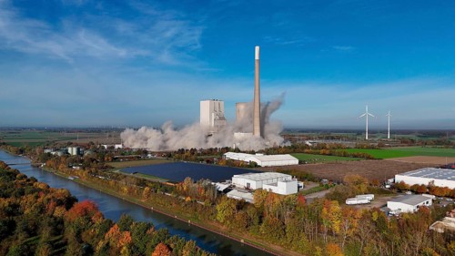 A power plant is being demolished, smoke rises, wind turbines stand next to it in an autumn