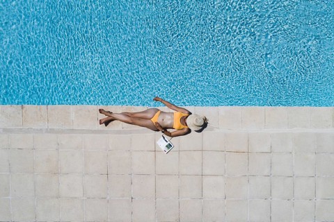 Young woman lying in the sun at the edge of a swimming pool, her face covered by a sun hat,