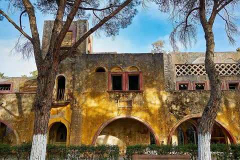 Ruined building with yellow façade and arches flanked by tall trees with blue sky, Italian