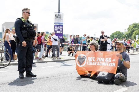 A police officer observes two Last Generation activists with a banner on Article 20A of the Basic