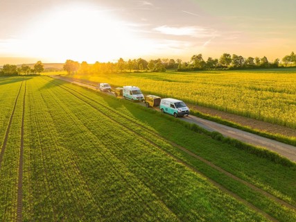 Cars driving along a rural road next to green fields at sunset, fibreglass installation, Calw