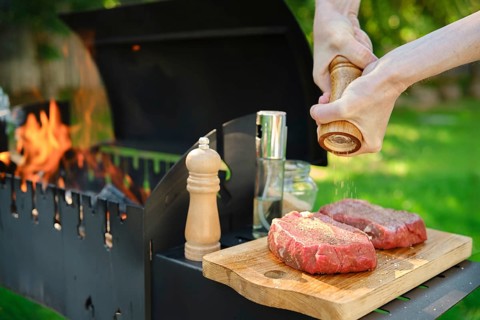 A close-up view of hands using a wooden hand mill to season beef steak with pepper