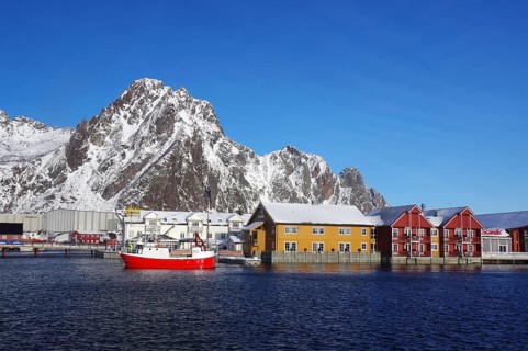 Colourful houses and a red fishing boat in the harbour in front of snow-covered mountains under a