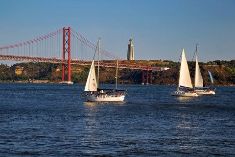 Two sailboats on calm waters with a red suspension bridge in the background under a blue sky,