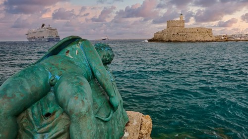 A damaged sculpture looks out over the sea and a fortress during a dramatic sunset, sky exchanged,
