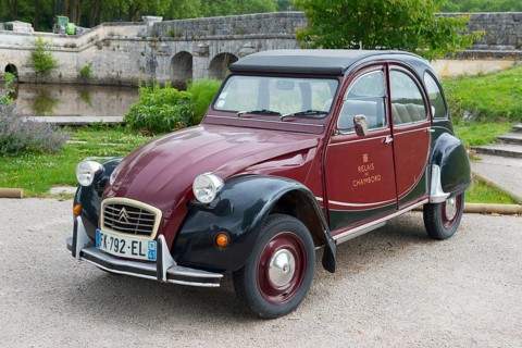 Retro Citroën 2CV vintage car in red and black, parked on a road in front of a stone bridge in a