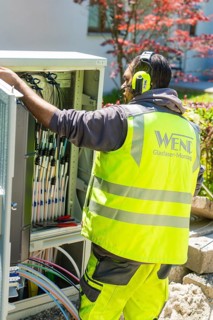 A worker in a safety waistcoat is busy with fibre optic assembly, surrounded by a garden with a red