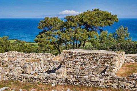 Ancient wall remains near a coast, surrounded by trees and a clear blue sky, the sea in the