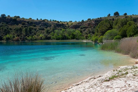 A calm lake with clear blue water, surrounded by green trees and rolling hills under a clear sky,
