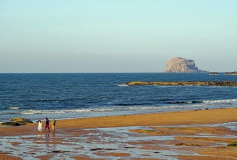 Walkers on the beach with rocks and waves in the background under a clear sky, Bass Rock, North