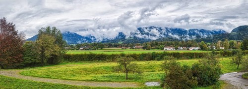Wide landscape with green meadows, trees and a village in front of a mountain range under a cloudy
