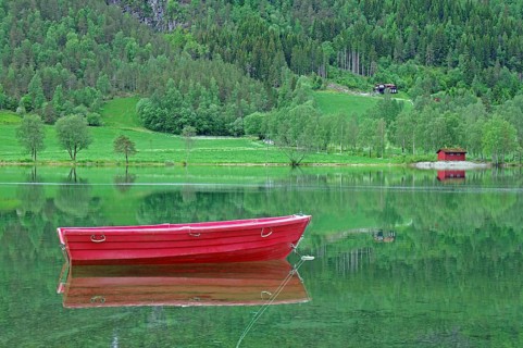 A small red boat floats on a calm lake with green surroundings, Stryn, Mindesundet, Fjordland,