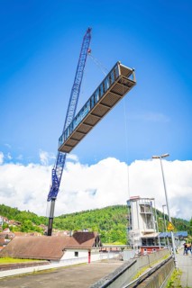 A crane lifts a bridge over a building complex in sunny weather with blue sky and white clouds,