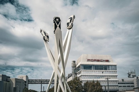 Modern sculpture of cars on high pillars in front of a modern building under a cloudy sky,