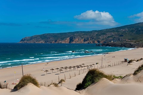 Beach with sand dunes and sunshades, behind them the sea and rocks, partly cloudy sky, moving dune,