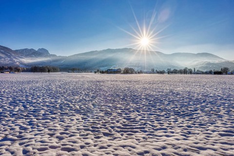 Winter landscape with snow-covered field and rising sun against a mountain backdrop, Bad Feilnbach