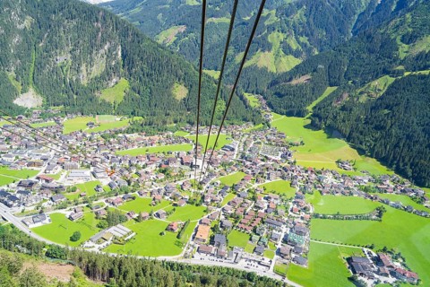 View from above of a town with surrounding mountains and green meadows, with cable car cable in the