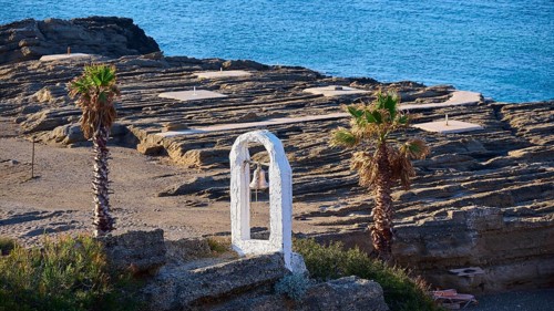 View of a rocky coastline with palm trees, a white bell frame and blue sky in the background, rock