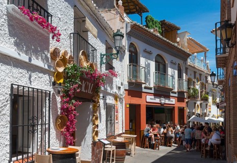 Narrow alley with white, ornate buildings and colourful floral decorations where people sit