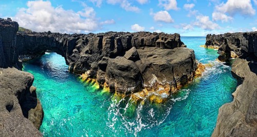 Rugged lava rock formations with a natural arch over a turquoise sea under a blue sky, A Porta do