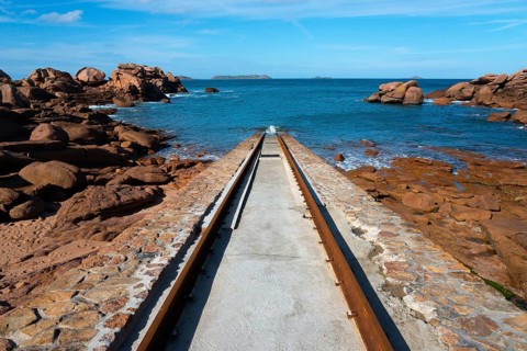 A stone path leads through a rocky coastal landscape to the blue sea under a clear sky, Cale de