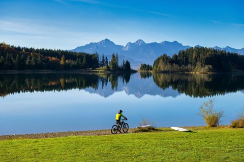 A cyclist rides along the shore of a lake, surrounded by mountains and reflecting trees in a clear