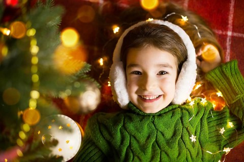 A happy girl in fur headphones lies near the Christmas tree in garlands and lights in a warm