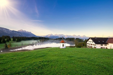 Farmhouse next to a chapel on a lake with a view of the Alps at sunrise, surrounded by green