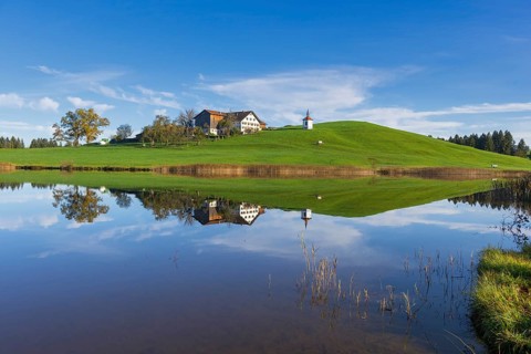View of a lake with reflection of a house and a chapel on a hill, Hegratsrieder See near Füssen,