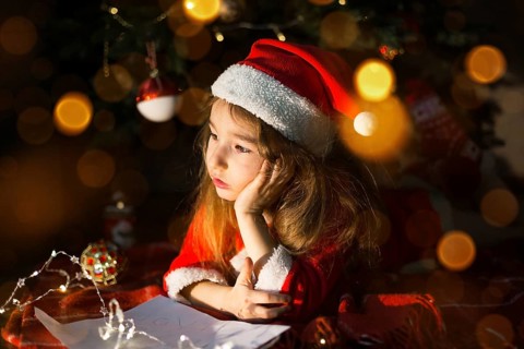 Little girl in a Santa hat and red dress under Christmas tree is dreaming, waiting for the holiday,