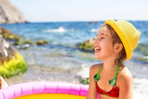 Girl in yellow straw hat plays in outdoor near sea, in water with a bucket in an inflatable pool on
