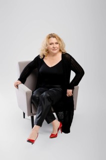 Happy senior woman in knitted cardigan and faux leather trousers sitting in armchair over grey