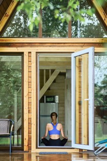 Back view of young woman sitting in lotus yoga pose inside wooden cabin in front of open door