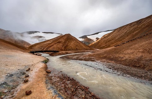 Bridge over steaming stream between colourful rhyolite mountains with snowfields, Hveradalir