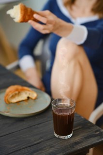 Unrecognizable woman sitting in the kitchen with croissant in hand, focus on glass with coffee