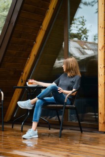 Woman sits in wicker chair near the table at wooden terrace of her forest cabin