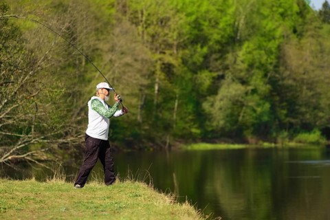 Fisherman with a spinning rod catching fish on a river at sunny summer day with green trees at