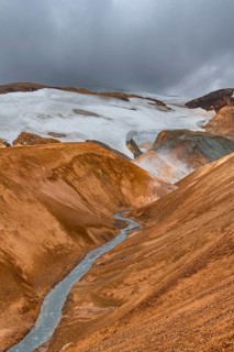 Steaming stream between colourful rhyolite mountains with snowfields, Hveradalir geothermal area,