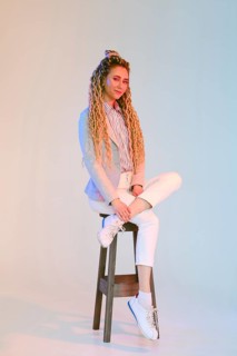Young woman with dreadlocks in jeans and jacket sits on chair under colorful lights